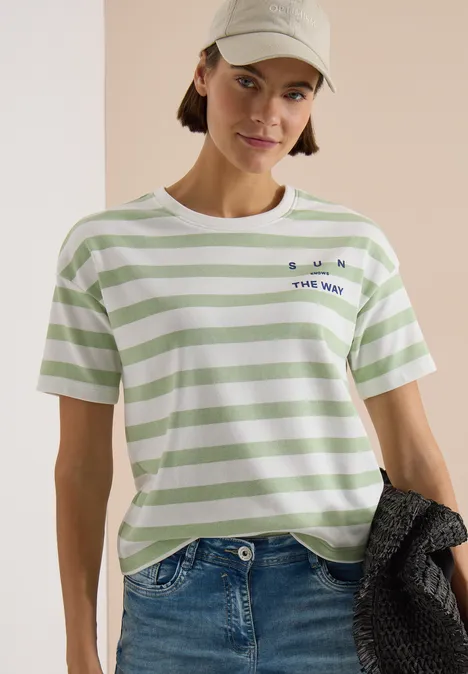 Vrouw in groen-wit gestreept T-shirt, jeans en pet, houdt een gebreid vest vast.