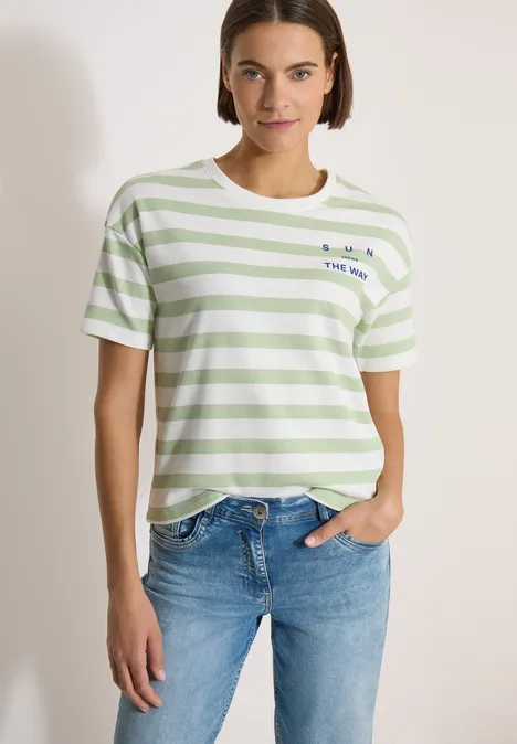 Vrouw met groen-wit gestreept T-shirt en blauwe jeans voor een witte muur.