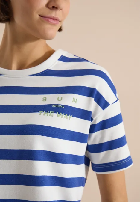 Vrouw draagt wit-blauw gestreept T-shirt met groene tekst 'SUN KNOWS THE WAY'.