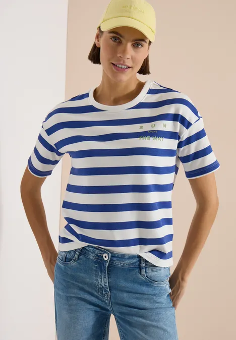 Persoon in wit-blauw gestreept T-shirt en spijkerbroek, handen in zij voor een neutrale achtergrond.