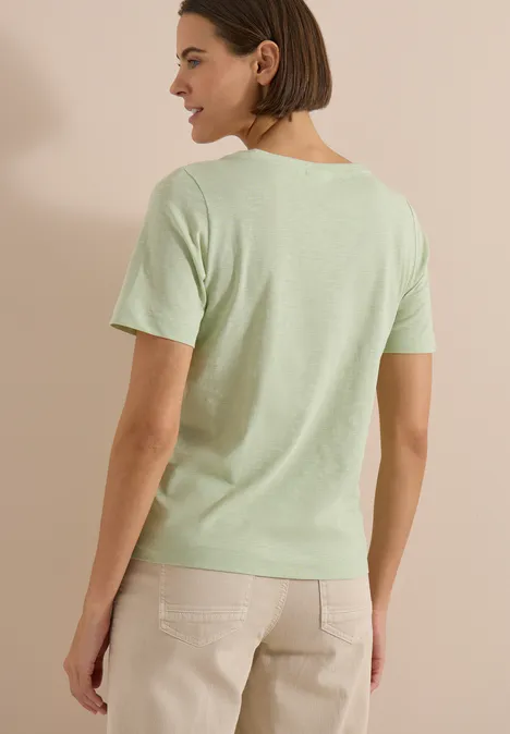 Rugaanzicht van een vrouw in een lichtgroen T-shirt en beige broek tegen neutrale achtergrond.