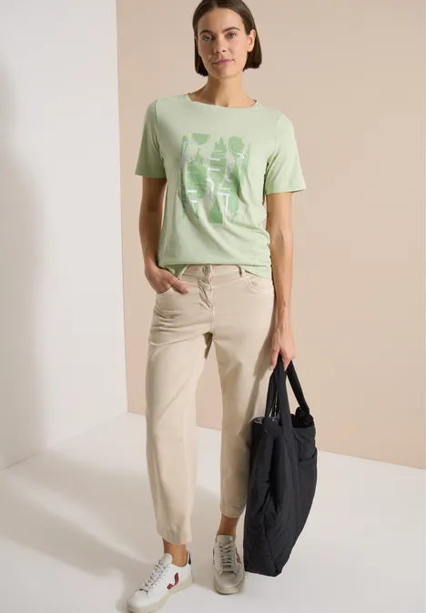 Vrouw in lichtgroen T-shirt met print, crème broek, witte sneakers en donkerblauwe tas.