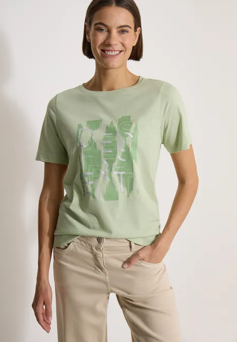 Vrouw in lichtgroen T-shirt met print en beige broek, staande met hand in zak.