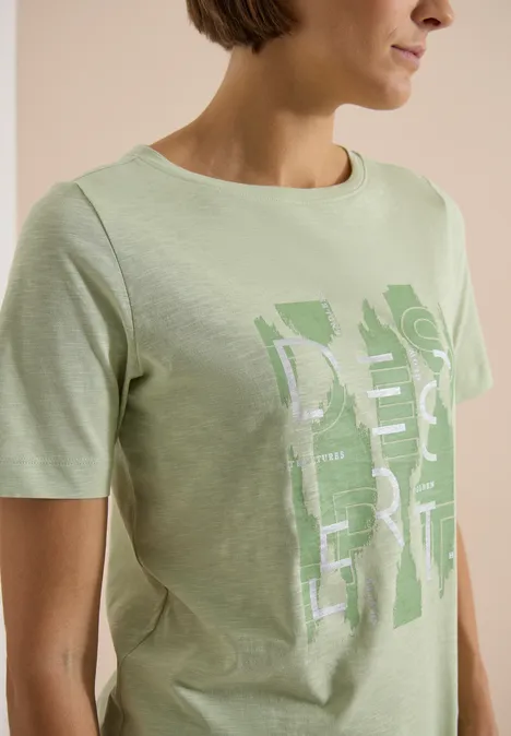 Vrouw in lichtgroen T-shirt met grafische witte/groene print, tegen een eenvoudige beige achtergrond.
