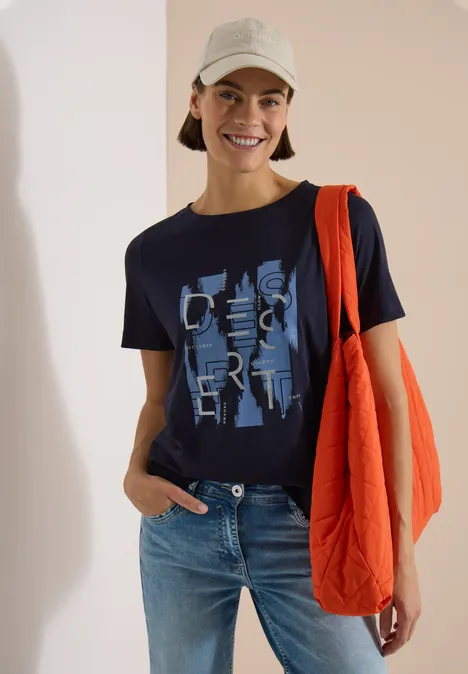 Shirt met ronde hals en opdruk aan de voorkant urban dark blue Persoon in jeans en donkerblauw shirt met print, draagt oranje tas en lichte bucket hat voor lichte achtergrond.