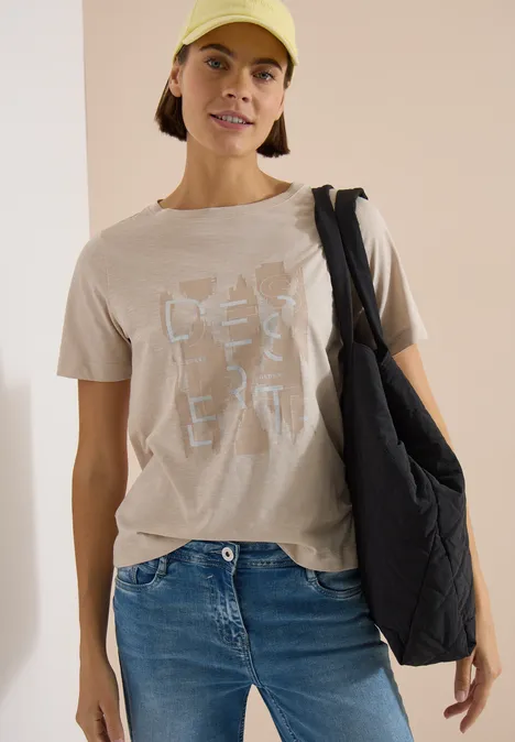 Vrouw draagt een beige T-shirt met witte print, een blauwe spijkerbroek en een zwarte gewatteerde schoudertas.