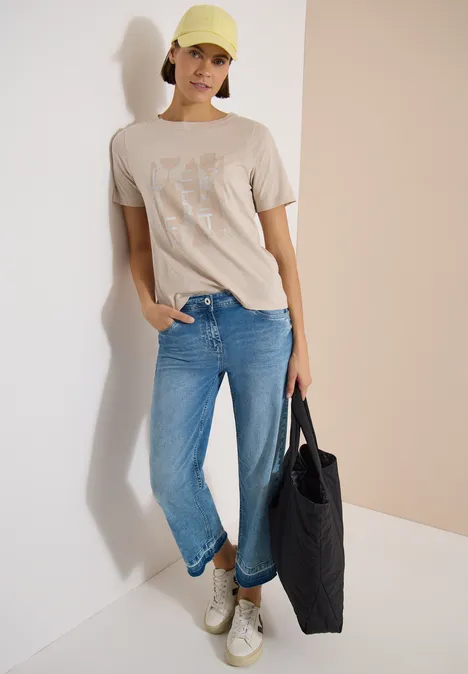 Persoon in beige T-shirt, blauwe spijkerbroek, witte sneakers en zwarte tas.