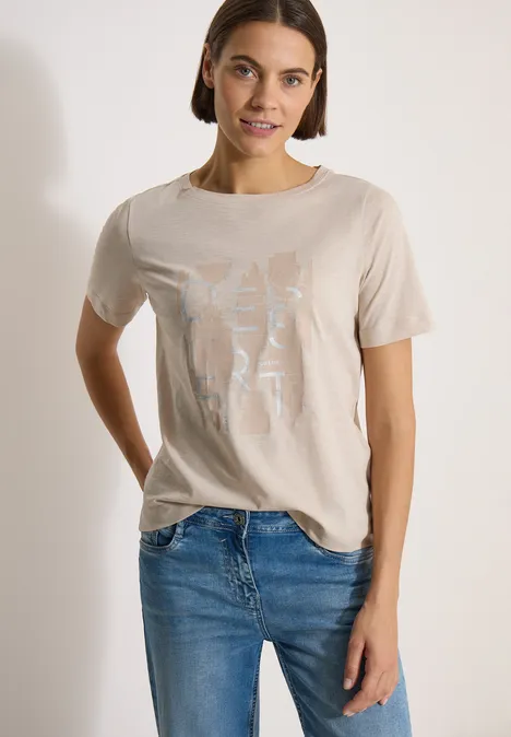 Vrouw in beige T-shirt met lichte print en blauwe jeans voor een witte achtergrond.