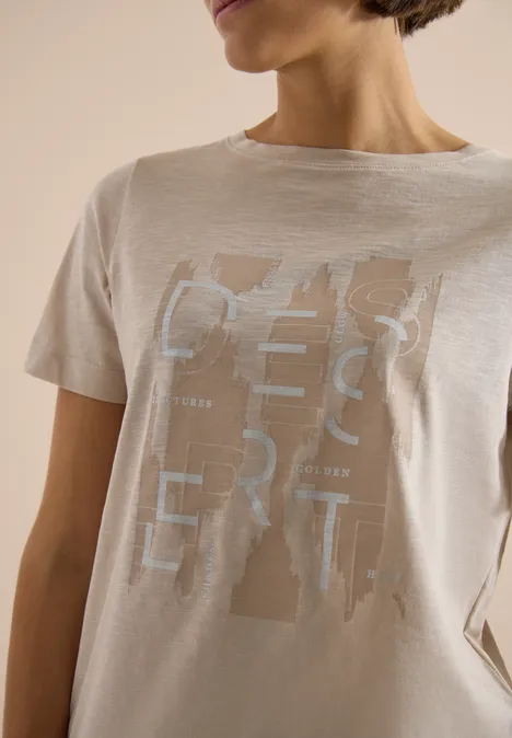 Vrouw draagt een beige t-shirt met abstracte zilverkleurige tekstprint