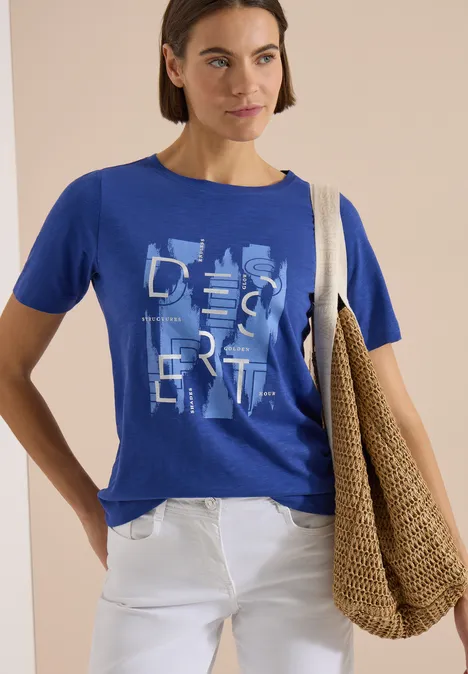 Vrouw draagt blauw T-shirt 'Desert', witte broek en grote strozak over de schouder