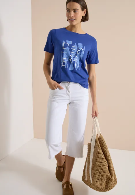 Vrouw in blauw T-shirt met print, witte cropped jeans, bruine loafers en grote gevlochten rieten tas.
