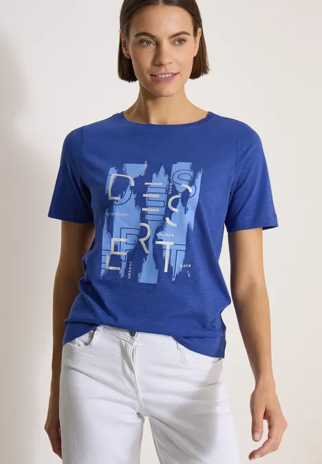 Persoon draagt blauw T-shirt met abstracte letters en witte broek, staand voor een lichte achtergrond.