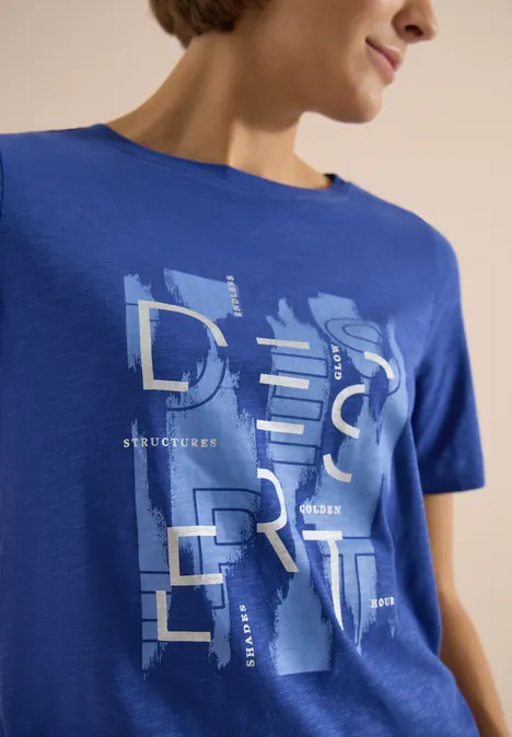 Vrouw draagt felblauw T-shirt met grafische, abstracte opdruk 'DESERT' voorop.