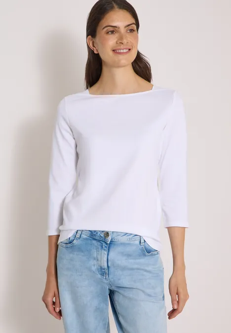 T-shirt basique à col bateau White Frau in weißem Dreiviertelarm-Shirt und hellblauen Jeans lächelt seitlich vor hellem Hintergrund.