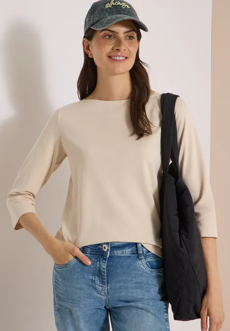 Frau mit cremefarbenem Oberteil, blauen Jeans, Baseballcap und schwarzer Tasche vor hellem Hintergrund.