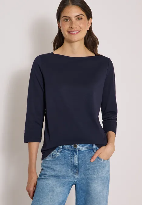 Frau mit schulterlangen braunen Haaren in marineblauem Oberteil und hellblauen Jeans, Hand in Hosentasche.