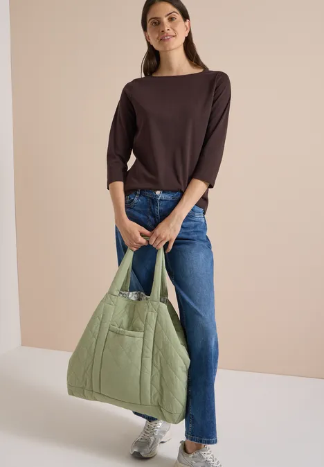 Frau in braunem Shirt und Jeans trägt eine hellgrüne, gesteppte Tasche und weiße Sneaker.