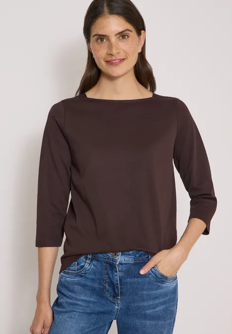 Frau in braunem Dreiviertelarm-Shirt und Jeans, Hände in Taschen, lächelnd vor hellem Hintergrund.