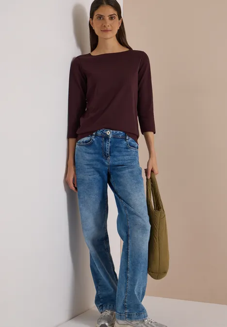 Basic Shirt met boothals berry red Frau steht vor einer Wand, trägt bordeauxfarbenes Shirt, blaue Jeans und eine grüne Tasche.