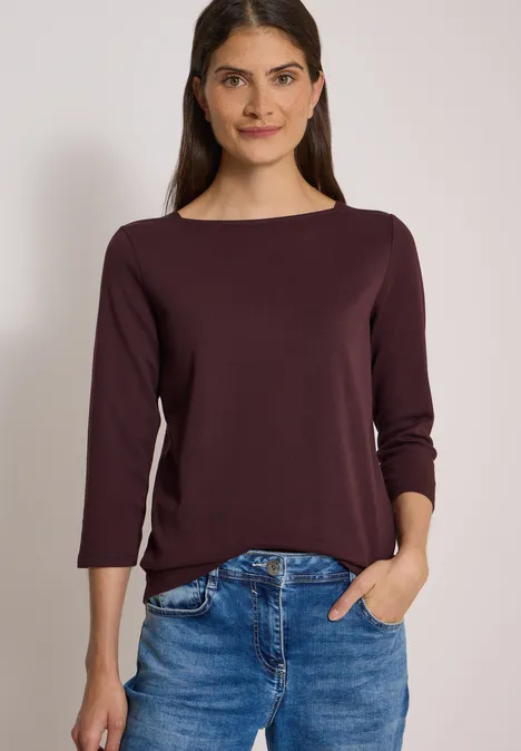 Basic Shirt met boothals berry red Eine lächelnde Frau in weinrotem Langarmshirt mit U-Boot-Ausschnitt und Jeans, eine Hand in der Hosentasche.