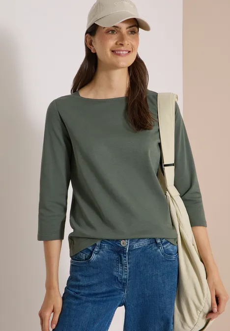 Frau in casual grünem 3/4-Shirt und Jeans, trägt eine Beuteltasche über der Schulter, lächelt.