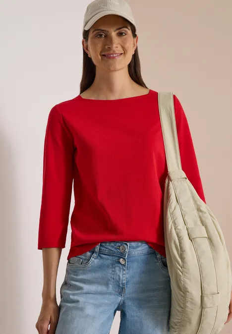 Basic shirt met boothals flame red Frau trägt rote Bluse mit 3/4-Ärmeln, Jeans und beige Stepp-Umhängetasche, lächelnd mit Baseballcap.