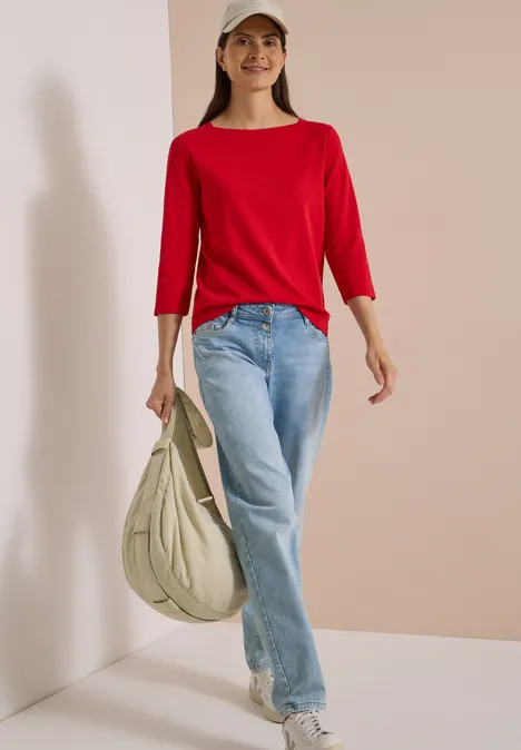 Basic shirt met boothals flame red Frau in rotem Shirt, Jeans, weißen Sneakern und Cap mit großer beiger Tasche vor neutralem Hintergrund.