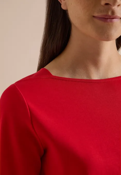 Basic shirt met boothals flame red Frau im roten Oberteil, Teil des Gesichts und Nacken zu sehen, braunes zurückgebundenes Haar.