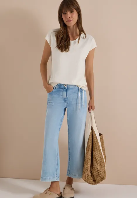 Frau in weißem T-Shirt, hellblauen, weiten Jeans und mit großer Strohtasche vor beigem Hintergrund.
