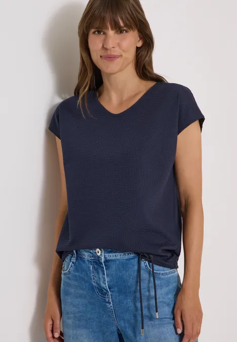 Frau trägt dunkelblaues T-Shirt mit kurzen Ärmeln und Jeans, lächelt leicht.