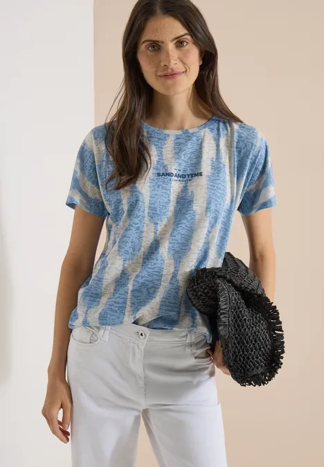 Shirt in uitgebleekte look met tekst urban dark blue Vrouw in blauw-wit T-shirt en witte broek met een zwarte tas voor een beige achtergrond.