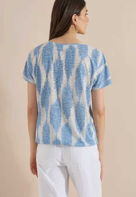 Shirt in uitgebleekte look met tekst urban dark blue Vrouw van achteren in een blauw-wit dessintrui met korte mouwen en witte broek tegen lichte achtergrond.