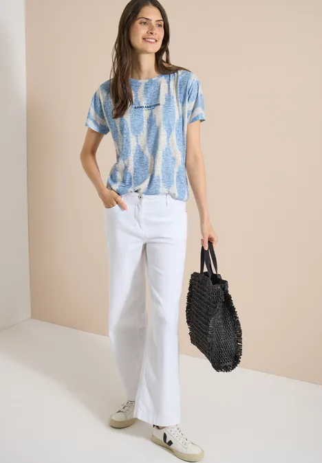 Shirt in uitgebleekte look met tekst urban dark blue Vrouw in blauw-wit shirt, witte jeans, sneakers en zwarte tas tegen neutrale achtergrond.
