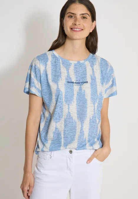 Shirt in uitgebleekte look met tekst urban dark blue Vrouw met blauw-wit T-shirt met dessin en witte jeans, voor een lichte achtergrond, hand in de zak.