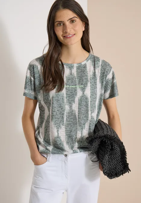 Shirt in uitgebleekte look met tekst deep matcha green Vrouw in kort mouw T-shirt met groen-grijs patroon, witte broek en ze houdt een zwart jasje vast.