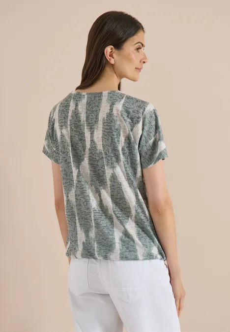 Shirt in uitgebleekte look met tekst deep matcha green Vrouw van achteren met los haar in een los vallende top met groen-wit patroon en een witte broek.