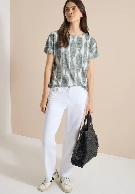 Shirt in uitgebleekte look met tekst deep matcha green Vrouw in witte jeans, slangenblouse en sneakers, met grote zwarte gevlochten tas tegen beige achtergrond.