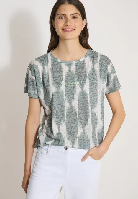 Shirt in uitgebleekte look met tekst deep matcha green Vrouw in grijs t-shirt met wit patroon en witte broek, staand met één hand in de zak.