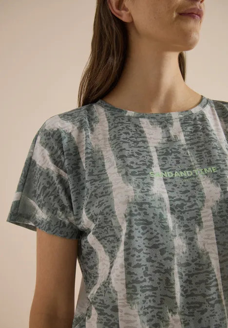 Shirt in uitgebleekte look met tekst deep matcha green Close-up van een vrouw met een grijs-groen T-shirt met abstract patroon en subtiele tekst.