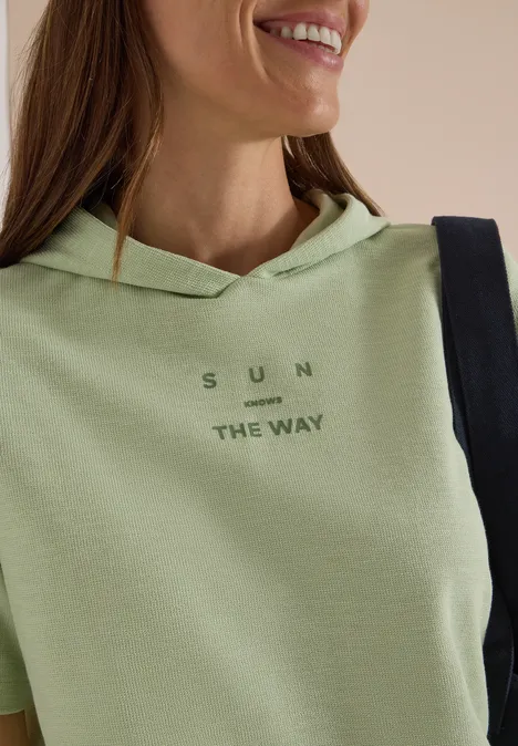 Kurzarm Shirt mit Kapuze und Wording raw matcha lime Frau mit hellem grünen Hoodie, Aufschrift 'SUN THE WAY', lächelt, schwarze Tasche über der Schulter.