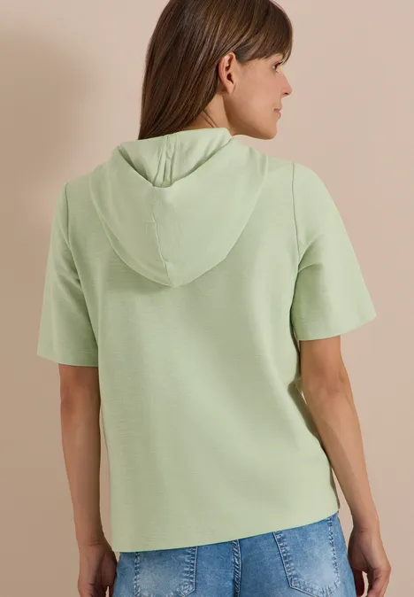 Kurzarm Shirt mit Kapuze und Wording raw matcha lime Frau von hinten in hellgrünem Kurzarm-Hoodie und blauen Jeans.
