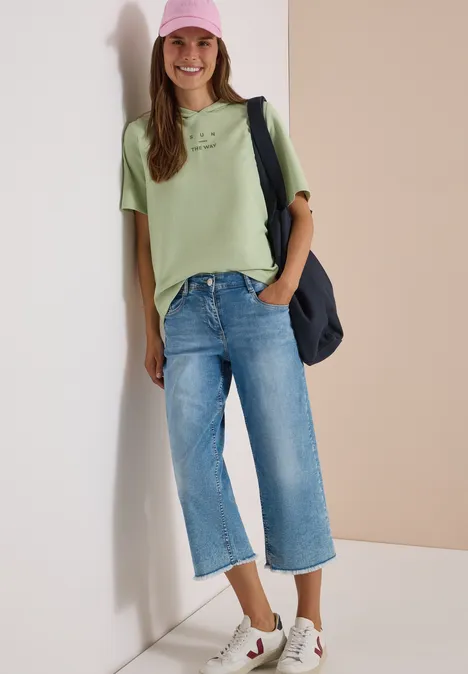 Kurzarm Shirt mit Kapuze und Wording raw matcha lime Junge Frau lehnt an Wand, trägt grünes T-Shirt, Jeans-Culotte, rosa Cap, Sneaker und Schultertasche.