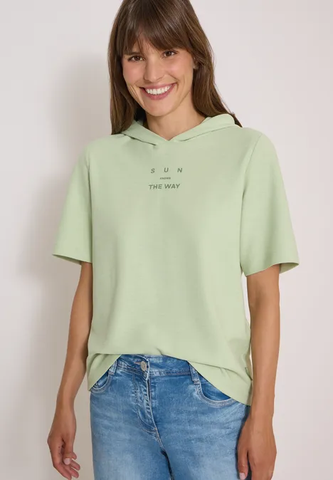 Kurzarm Shirt mit Kapuze und Wording raw matcha lime Junge Frau lächelt, trägt ein hellgrünes, kurzärmeliges Kapuzenshirt und blaue Jeans vor hellem Hintergrund.