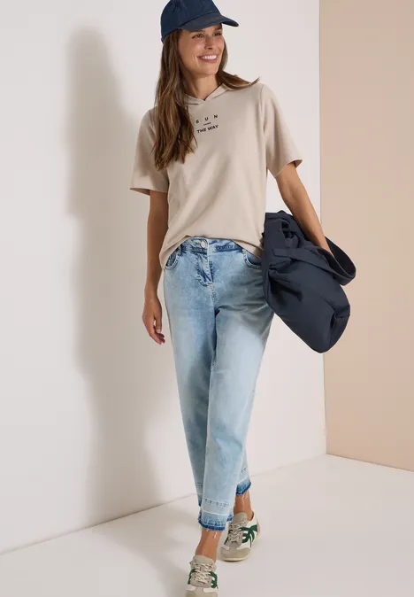 Kurzarm Shirt mit Kapuze und Wording cashmere beige Junge Frau in beige T-Shirt, hellen Jeans und Cap, mit Jacke über dem Arm, lächelnd vor weißer Wand.