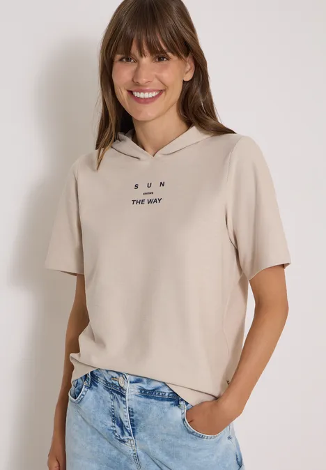 Kurzarm Shirt mit Kapuze und Wording cashmere beige Junge Frau lächelt, trägt beiges, kurzärmliges Kapuzen-Shirt mit Aufdruck und Jeans.