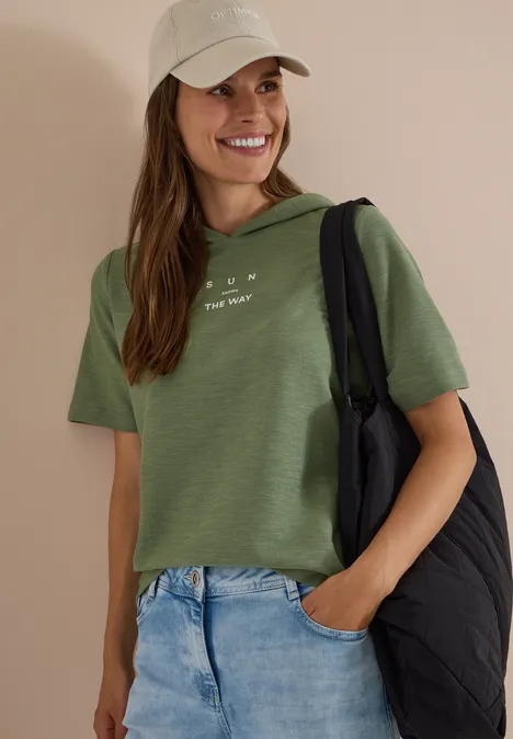 Kurzarm Shirt mit Kapuze und Wording deep matcha green Eine lächelnde Frau in grüner Bluse, Jeans und Cap mit schwarzer gesteppter Schultertasche.