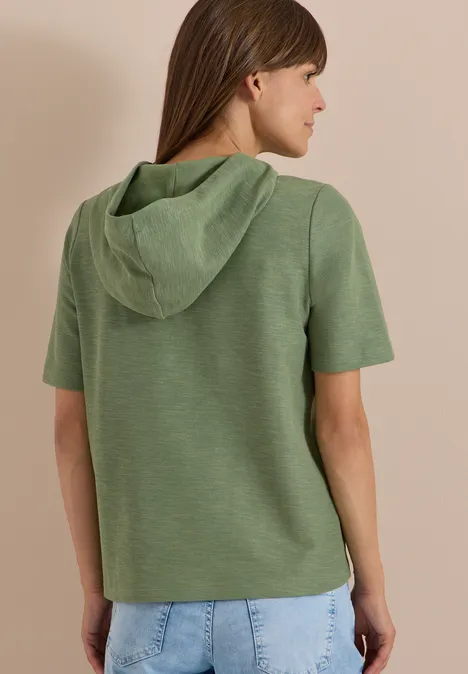 Kurzarm Shirt mit Kapuze und Wording deep matcha green Eine Frau von hinten in grünem Kurzarm-Hoodie und hellblauen Jeans vor hellem Hintergrund.