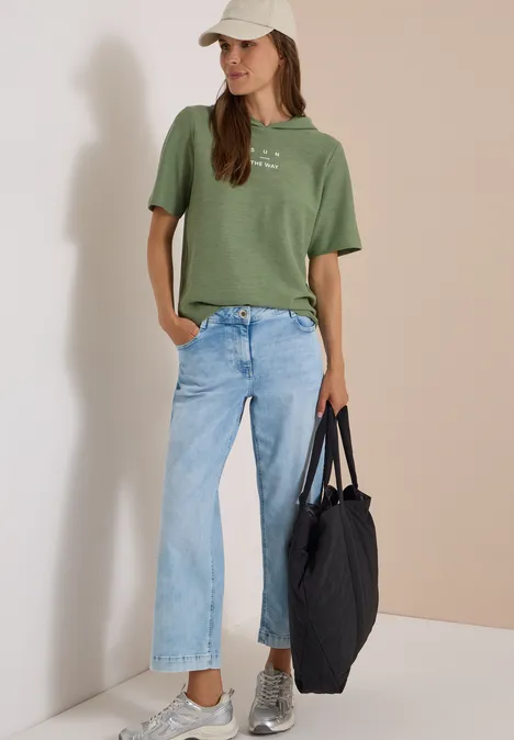 Kurzarm Shirt mit Kapuze und Wording deep matcha green Frau in grünem T-Shirt, hellen Jeans, grauen Sneakern, weißer Cap und schwarzer Tasche vor schlichter Wand