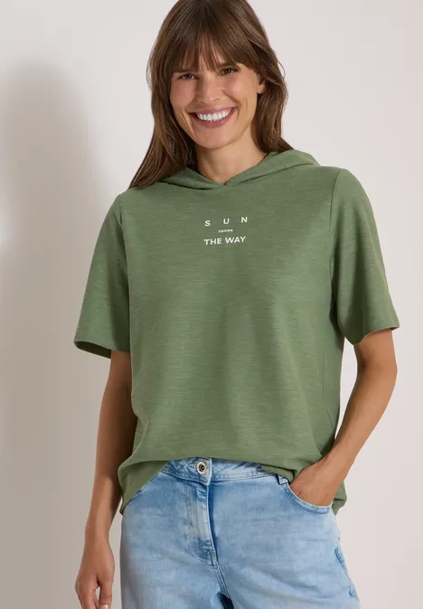 Kurzarm Shirt mit Kapuze und Wording deep matcha green Frau mit kurzem, grünem Kapuzen-Shirt und hellblauen Jeans, lächelnd mit Händen in den Taschen.