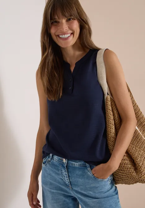 Lächelnde Frau mit braunen Haaren, blauem ärmellosen Top, Jeans und großer gewebter Strandtasche.